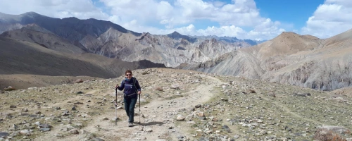 Trekker walking on a rocky mountain trail with barren hills and peaks in the background.