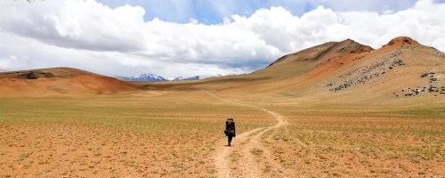 Trekker walking along a winding trail across a vast barren valley with distant hills.