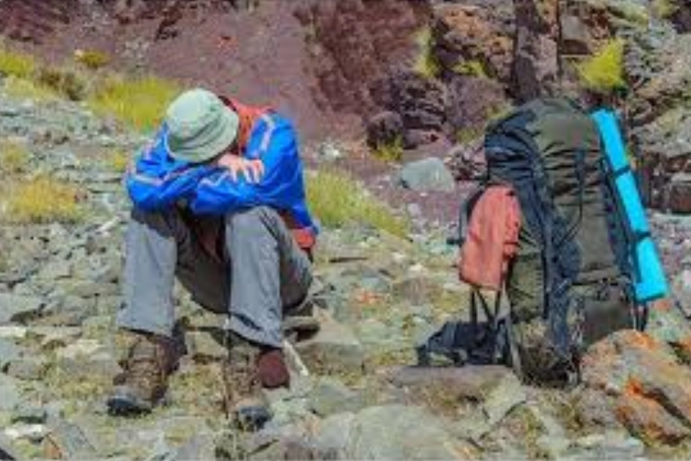 Trekker resting on a rocky mountain trail beside a large backpack during a high-altitude hike.