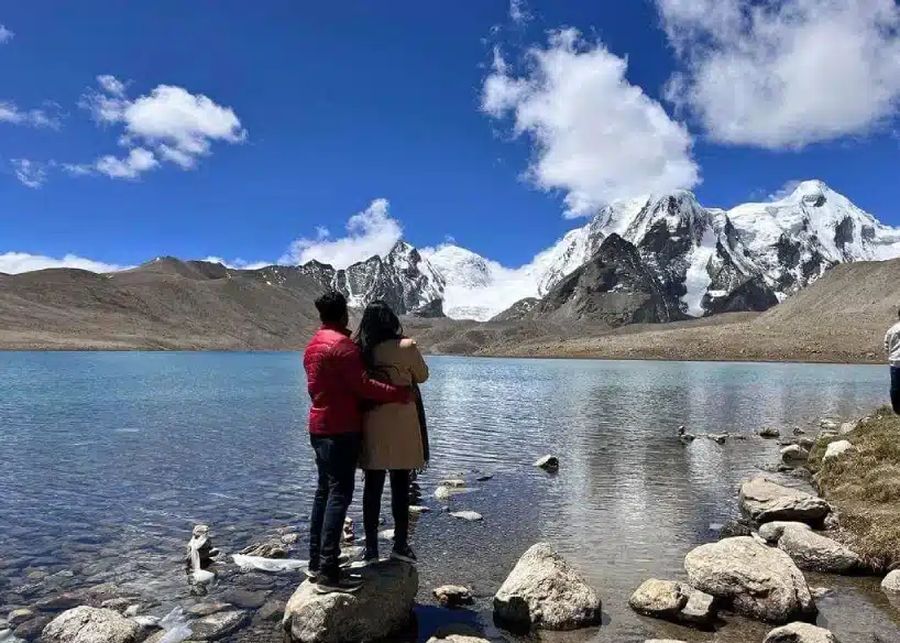Couple standing by a serene mountain lake with snow-covered Himalayan peaks in Sikkim.